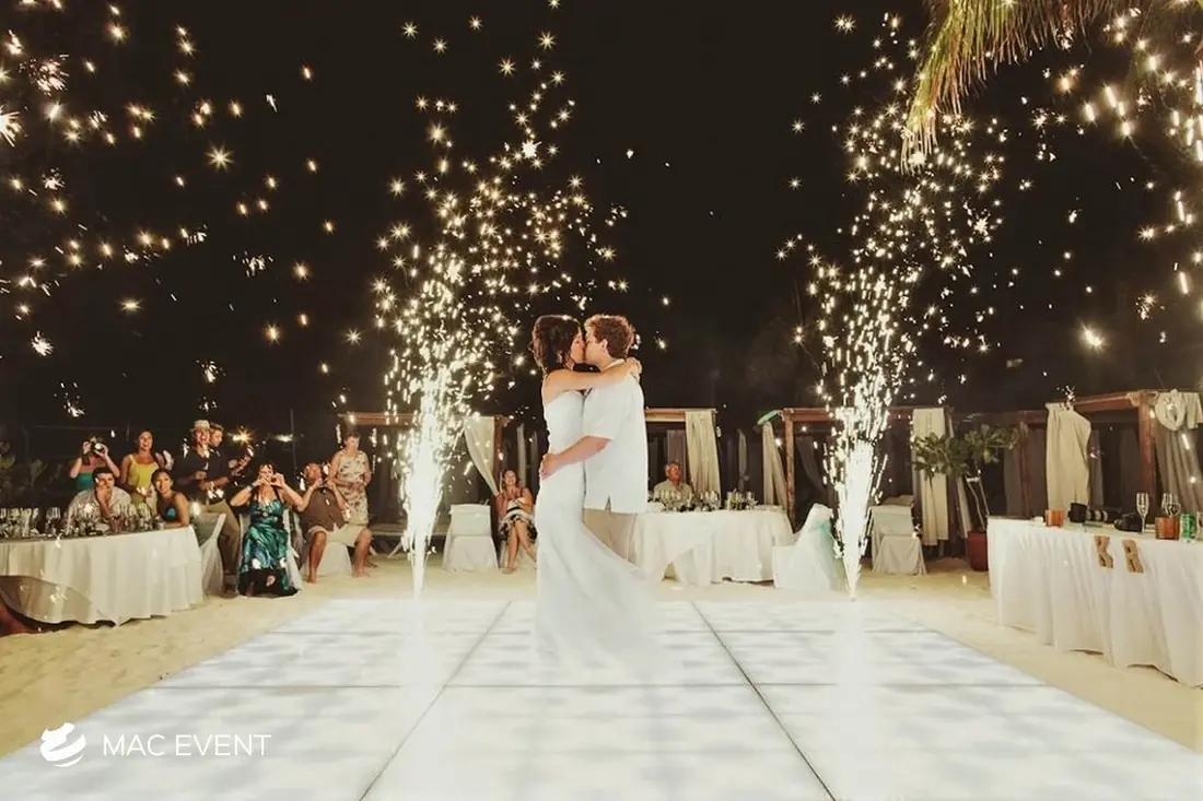 A newlywed couple embraces on a white dance floor, surrounded by sparkling fireworks and cheering guests at a tropical beachside wedding reception.