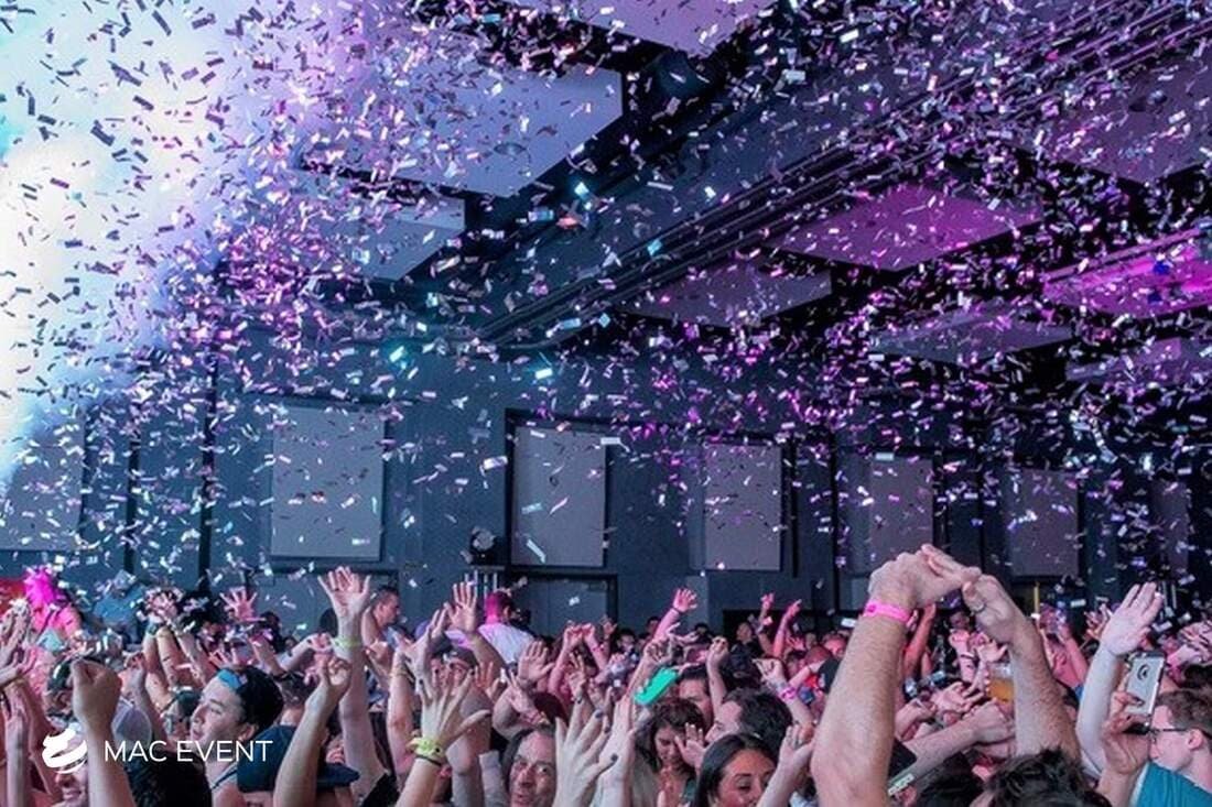 A high-energy party scene with a crowd dancing under a confetti shower, illuminated by purple and blue stage lighting in a nightclub-like venue.