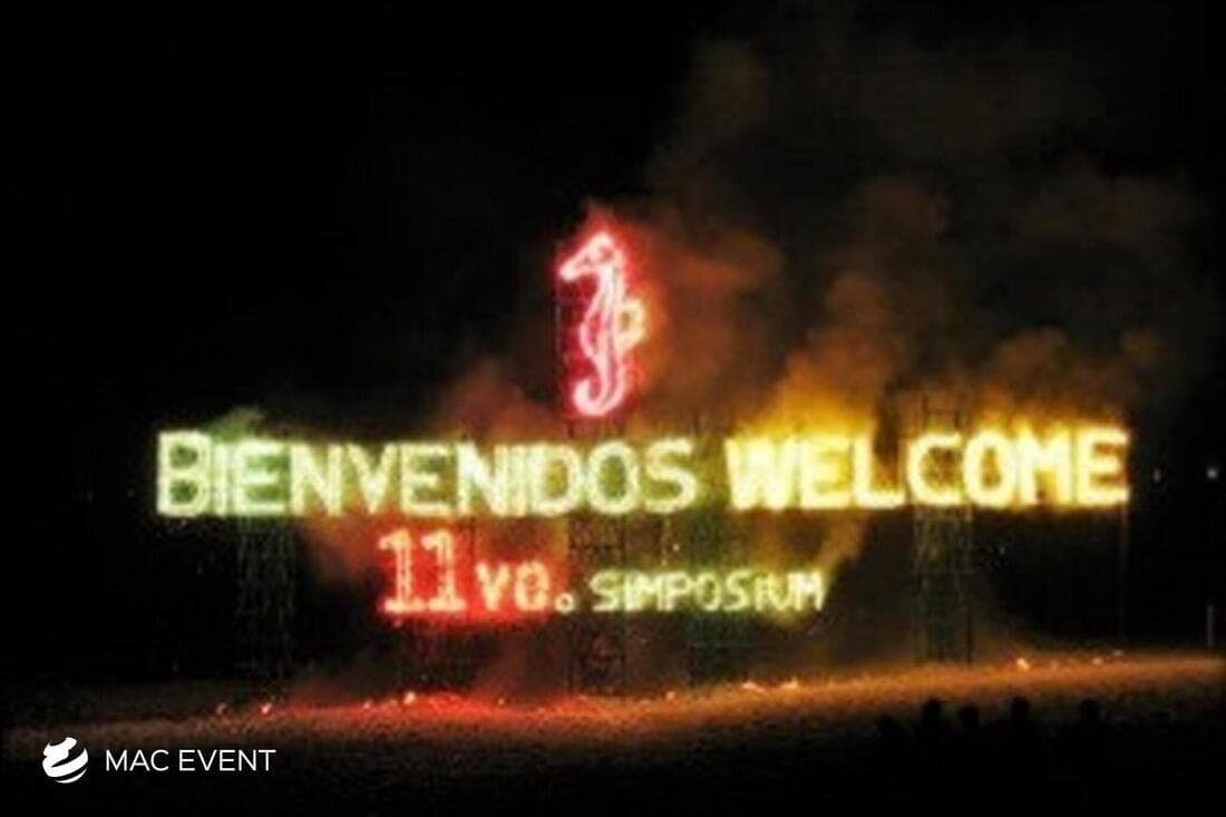 A fire-lit welcome sign at an event, spelling "Bienvenidos Welcome" alongside a red seahorse symbol and the words "11vo Simposium," glowing against a smoky background.