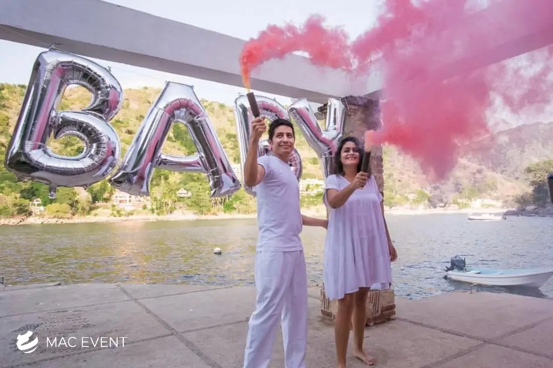 A couple dressed in white celebrates a gender reveal by holding pink smoke bombs, with large silver balloons spelling "BABY" in the background.