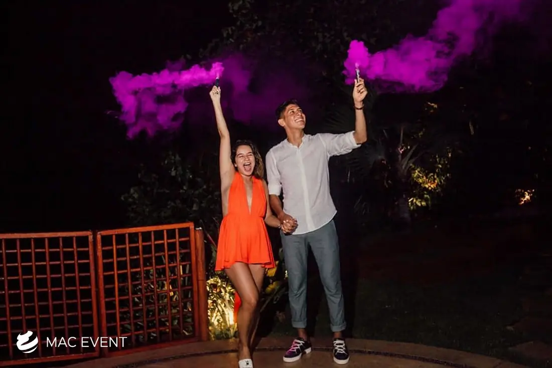 A joyful couple at an outdoor nighttime event waves purple smoke bombs in the air while holding hands and celebrating with excitement.